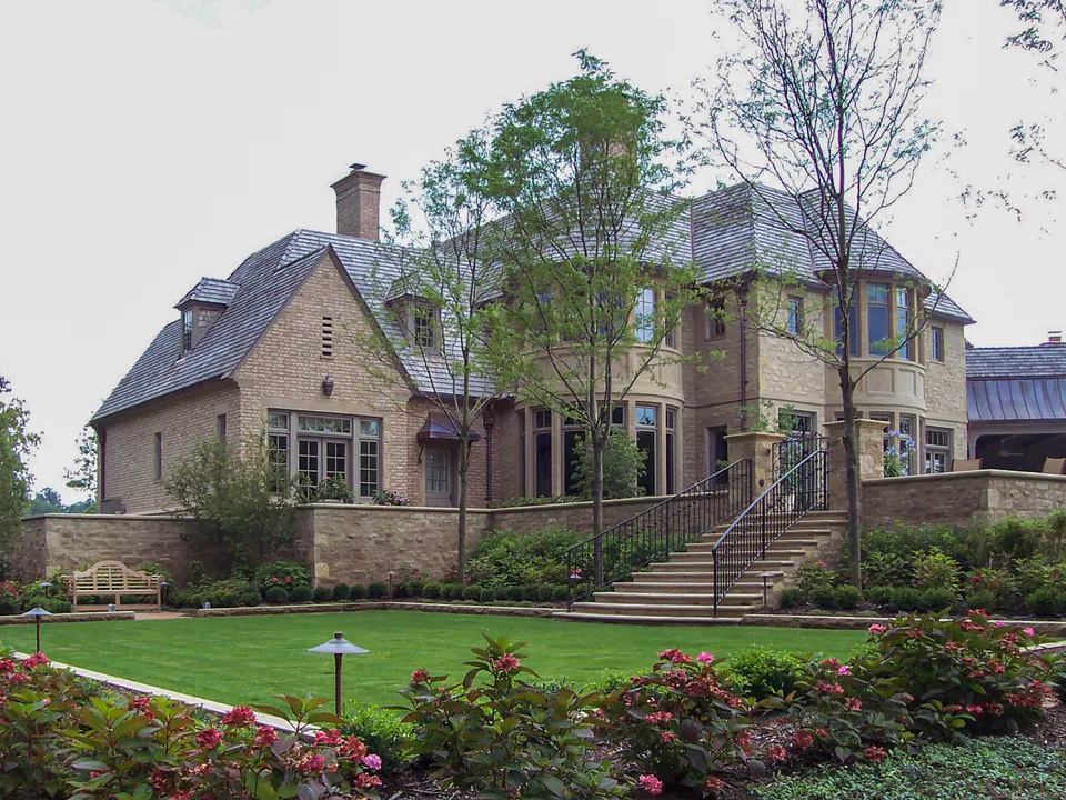Rear estate lawn with stone house, broad stairs, railings, stone walls, benches, and layered planting.